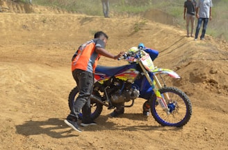 A person appears to be assisting a motorcyclist in an off-road sandy terrain. The motorbike is decorated with vibrant colors and the rider is wearing a helmet and racing gear. In the background, a couple of individuals are standing, observing the scene.