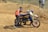 A person appears to be assisting a motorcyclist in an off-road sandy terrain. The motorbike is decorated with vibrant colors and the rider is wearing a helmet and racing gear. In the background, a couple of individuals are standing, observing the scene.