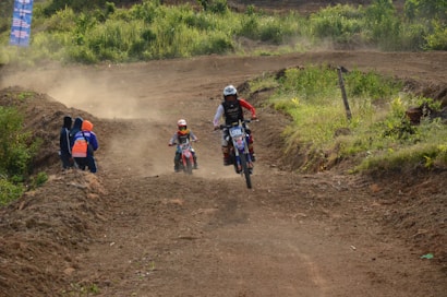 A dirt bike race is taking place on a rugged dirt track, surrounded by green vegetation. Two riders wearing helmets and protective gear are actively maneuvering their bikes on the course. A group of people stands off to the side of the track, observing the race. Dust clouds rise from the bikes, suggesting high speed and dynamic movement.