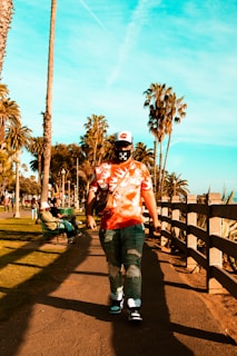 A person wearing a tie-dye shirt, jeans, and a cap with a distinct pattern is walking along a sunlit path lined with palm trees. The sky is bright blue, providing a vivid backdrop. People are seen sitting on benches and enjoying the outdoors. The scene conveys a leisurely and relaxed atmosphere.