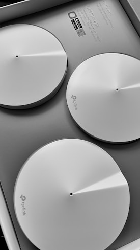 Three circular, white devices with a minimalist design are placed in a neatly organized manner inside a box. Each device has a small hole in the center and a subtle logo of 'TP-Link'. The box includes instructions and QR codes for downloading an app, suggesting they are likely part of a tech product setup.