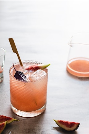 An elegant cocktail glass filled with a deep red drink, surrounded by fresh figs and herbs.