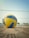 A volleyball lies on a sandy court in front of a net. The ball is prominently positioned in the foreground, displaying its yellow and blue panels. In the background, there is a clear blue sky and a grassy area with some fencing and structures.