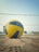 A volleyball lies on a sandy court in front of a net. The ball is prominently positioned in the foreground, displaying its yellow and blue panels. In the background, there is a clear blue sky and a grassy area with some fencing and structures.