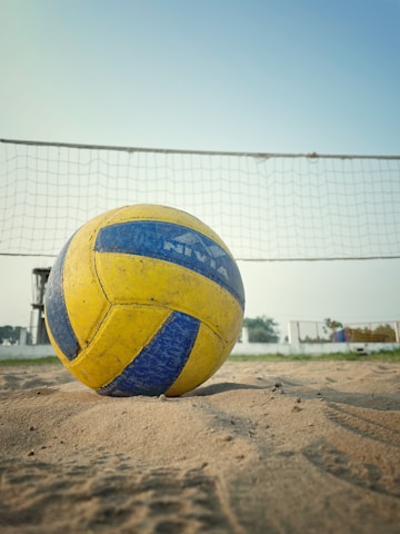 A volleyball lies on a sandy court in front of a net. The ball is prominently positioned in the foreground, displaying its yellow and blue panels. In the background, there is a clear blue sky and a grassy area with some fencing and structures.