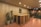 A modern reception area features a wooden desk with vertical panels and decorative objects on top, including a small sculpture and a potted plant. Two large potted plants with broad leaves are placed beside the desk. The ceiling is equipped with recessed lighting, and the floor is tiled with red terracotta tiles. The space extends into a corridor with additional plants and a wooden structure visible.