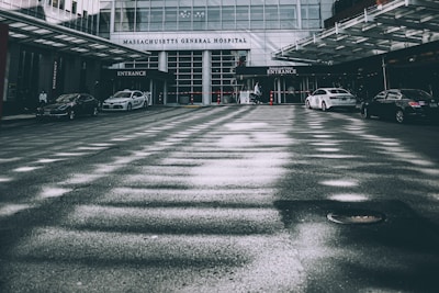 A hospital entrance is visible with multiple cars parked in front of it. The building is modern with glass windows and a prominent 'Massachusetts General Hospital' sign. A few people can be seen near the entrance, and the overall setting appears to be calm with an overcast lighting casting shadows on the pavement.