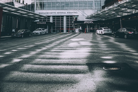 A hospital entrance is visible with multiple cars parked in front of it. The building is modern with glass windows and a prominent 'Massachusetts General Hospital' sign. A few people can be seen near the entrance, and the overall setting appears to be calm with an overcast lighting casting shadows on the pavement.