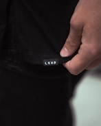 A close-up image of a person's hand holding the hem of a black garment with a small white label displaying the letters 'LSKD'. The lighting is soft, creating subtle shadows and highlights on the fabric and skin.