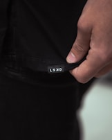 A close-up image of a person's hand holding the hem of a black garment with a small white label displaying the letters 'LSKD'. The lighting is soft, creating subtle shadows and highlights on the fabric and skin.