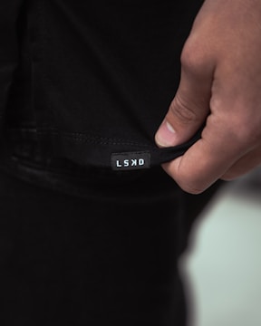 A close-up image of a person's hand holding the hem of a black garment with a small white label displaying the letters 'LSKD'. The lighting is soft, creating subtle shadows and highlights on the fabric and skin.