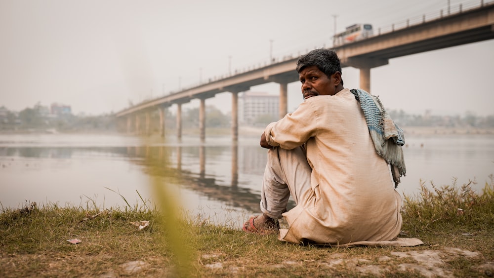 man in brown coat sitting on green grass near body of water during daytime