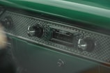 A vintage car dashboard featuring an old-style radio with a band and auxiliary interface. The dashboard has a textured surface with metallic knobs for volume and tuning. The dominant color is a rich green, giving it a retro aesthetic.