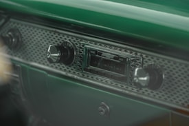 A vintage car dashboard featuring an old-style radio with a band and auxiliary interface. The dashboard has a textured surface with metallic knobs for volume and tuning. The dominant color is a rich green, giving it a retro aesthetic.