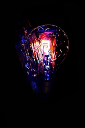 A clear light bulb with visible filaments is illuminated against a dark background. Vibrant colors of blue, purple, and red create a luminous effect, highlighting the intricate structure of the bulb and its internal details.