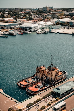 Two tugboats are docked in a harbor with clear blue water. In the background, there is a view of a coastal town with a variety of buildings, including residential houses with white and pastel-colored roofs. A few people are walking along the waterfront near greenery and a parked bus.