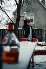 A person in a black jacket and orange cap is playing a game of beer pong outdoors in the snow. The table is set with red cups arranged for the game. Snow is falling, and a bottle is blurred in the foreground. The setting includes a wooden fence and a house in the background. Bare tree branches add to the wintry atmosphere.