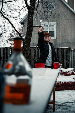 A person in a black jacket and orange cap is playing a game of beer pong outdoors in the snow. The table is set with red cups arranged for the game. Snow is falling, and a bottle is blurred in the foreground. The setting includes a wooden fence and a house in the background. Bare tree branches add to the wintry atmosphere.