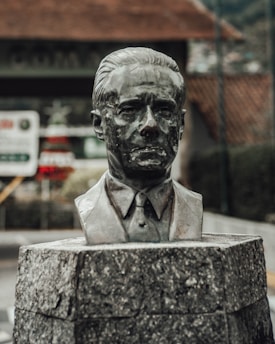 A bronze bust sculpture of a man in a suit is mounted on a textured stone pedestal. The background is blurred, showing hints of outdoor surroundings like a building and vegetation.