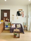 A cozy, well-decorated interior space featuring a small couch with patterned cushions covered by a plaid cloth. On the wall above, there are hanging clothes and decorative items, including a calendar displaying '2021' and a macrame piece. A small wooden table holds a vase with dried flowers and books beneath it. A bookshelf contains plants and decor. Soft natural light filters through a nearby window, creating a warm atmosphere.