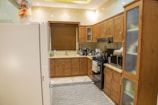 A modern kitchen with wooden cabinets, a white refrigerator, and a variety of kitchen appliances. There are flowers placed on top of the refrigerator, a window with a wooden blind above the sink, and an electric stove with a range hood. Various kitchen items including bottles and gadgets are placed on the countertops. The floor has a light-colored tile with a patterned rug.