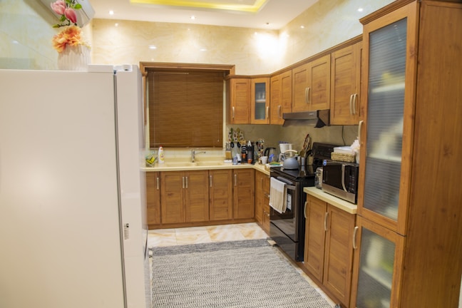 A modern kitchen with wooden cabinets, a white refrigerator, and a variety of kitchen appliances. There are flowers placed on top of the refrigerator, a window with a wooden blind above the sink, and an electric stove with a range hood. Various kitchen items including bottles and gadgets are placed on the countertops. The floor has a light-colored tile with a patterned rug.