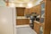 A modern kitchen with wooden cabinets, a white refrigerator, and a variety of kitchen appliances. There are flowers placed on top of the refrigerator, a window with a wooden blind above the sink, and an electric stove with a range hood. Various kitchen items including bottles and gadgets are placed on the countertops. The floor has a light-colored tile with a patterned rug.