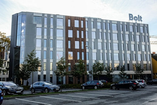 A modern office building with large windows and a mix of metallic and wood facade elements. There is a sign on top of the building that reads 'Bolt'. Several cars are parked in front of the building in a parking lot, and there are small trees lining the area between the parking spaces and the building. The sky is partly cloudy and the overall scene appears clean and orderly.