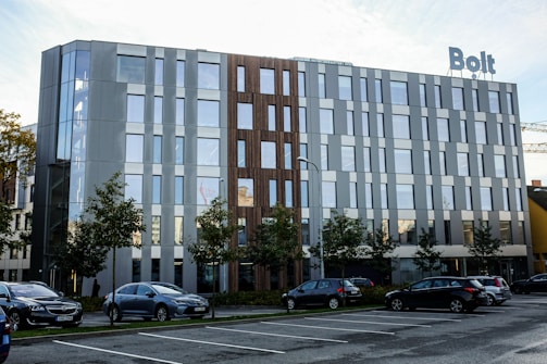 A modern office building with large windows and a mix of metallic and wood facade elements. There is a sign on top of the building that reads 'Bolt'. Several cars are parked in front of the building in a parking lot, and there are small trees lining the area between the parking spaces and the building. The sky is partly cloudy and the overall scene appears clean and orderly.