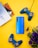 A blue smartphone is placed on a bright yellow background, surrounded by two blue game controllers with yellow buttons. To the side, there is a stack of colorful books, and a bunch of purple flowers adds a pop of color.
