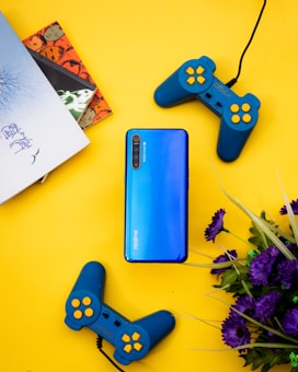 A blue smartphone is placed on a bright yellow background, surrounded by two blue game controllers with yellow buttons. To the side, there is a stack of colorful books, and a bunch of purple flowers adds a pop of color.