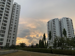 Two tall residential buildings are set against a partly cloudy sky with a sunset creating a warm, golden glow. Greenery, including trees and bushes, surrounds the buildings, with a well-maintained lawn in the foreground.
