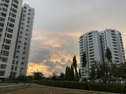 Two tall residential buildings are set against a partly cloudy sky with a sunset creating a warm, golden glow. Greenery, including trees and bushes, surrounds the buildings, with a well-maintained lawn in the foreground.
