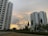 Two tall residential buildings are set against a partly cloudy sky with a sunset creating a warm, golden glow. Greenery, including trees and bushes, surrounds the buildings, with a well-maintained lawn in the foreground.