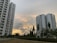 Two tall residential buildings are set against a partly cloudy sky with a sunset creating a warm, golden glow. Greenery, including trees and bushes, surrounds the buildings, with a well-maintained lawn in the foreground.
