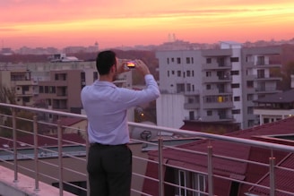 A person stands on a rooftop terrace, taking a photo with a smartphone. The viewpoint is elevated, overlooking a cityscape filled with modern apartment buildings. The sky is filled with vibrant hues of orange and pink, suggesting a sunset or sunrise, casting a warm glow over the scene.