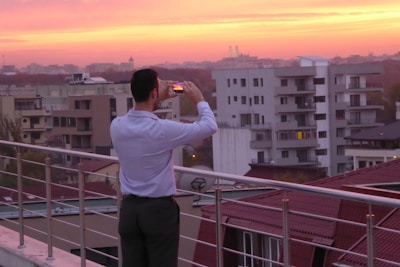 A person stands on a rooftop terrace, taking a photo with a smartphone. The viewpoint is elevated, overlooking a cityscape filled with modern apartment buildings. The sky is filled with vibrant hues of orange and pink, suggesting a sunset or sunrise, casting a warm glow over the scene.
