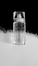 A clear, intricately designed perfume bottle is placed against a split black and white background. The bottle has a pattern resembling roses and a metallic label near the top. The lighting creates reflections, giving the bottle a pristine and elegant appearance.