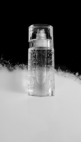 A clear, intricately designed perfume bottle is placed against a split black and white background. The bottle has a pattern resembling roses and a metallic label near the top. The lighting creates reflections, giving the bottle a pristine and elegant appearance.