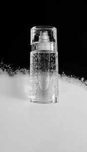 A clear, intricately designed perfume bottle is placed against a split black and white background. The bottle has a pattern resembling roses and a metallic label near the top. The lighting creates reflections, giving the bottle a pristine and elegant appearance.