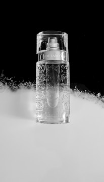 A clear, intricately designed perfume bottle is placed against a split black and white background. The bottle has a pattern resembling roses and a metallic label near the top. The lighting creates reflections, giving the bottle a pristine and elegant appearance.