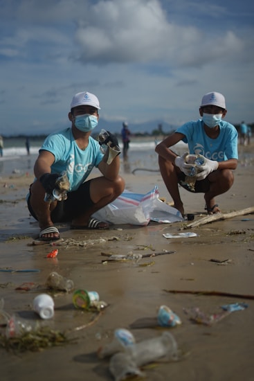Two individuals are crouched on a beach wearing masks, gloves, and matching light blue shirts with a logo. They appear to be participating in a cleanup effort, collecting plastic waste and other debris from the sand. A large white bag is placed nearby for collecting trash. The sky is partially cloudy with visible waves in the background.