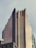 A tall, modern skyscraper with a unique, terraced glass facade reflecting the light. The building has multiple tiers that appear to taper as they ascend. The lower part of the image shows the rooftop of another building with a green trim. The sky is overcast, giving the scene a muted color palette.