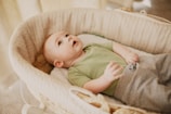 A baby is lying in a woven bassinet, looking upwards. The baby is wearing a green t-shirt and grey pants. The soft lighting in the room creates a warm and gentle atmosphere.