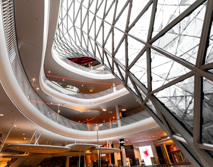 An abstract architectural interior featuring a modern, spiraling design with several levels. The ceiling is composed of a geometric pattern of glass and metal, letting in ample light and creating reflections. The structure blends curves and lines in a dynamic fashion, and the interior spaces feature warm lighting and open areas.