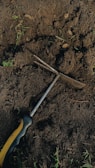 A hand tool with a black and yellow handle lies on dark, rich soil. The soil is slightly disturbed, suggesting digging or gardening activity. Small green sprouts and sticks are scattered around the area.