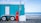 A turquoise trailer with white doors and wheels is parked on a rocky beach. A red propane gas cylinder and a metal pole with a sign are placed in front of the trailer. In the background, the ocean waves gently roll onto the shore under a partly cloudy sky.
