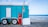 A turquoise trailer with white doors and wheels is parked on a rocky beach. A red propane gas cylinder and a metal pole with a sign are placed in front of the trailer. In the background, the ocean waves gently roll onto the shore under a partly cloudy sky.