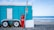 A turquoise trailer with white doors and wheels is parked on a rocky beach. A red propane gas cylinder and a metal pole with a sign are placed in front of the trailer. In the background, the ocean waves gently roll onto the shore under a partly cloudy sky.