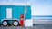 A turquoise trailer with white doors and wheels is parked on a rocky beach. A red propane gas cylinder and a metal pole with a sign are placed in front of the trailer. In the background, the ocean waves gently roll onto the shore under a partly cloudy sky.